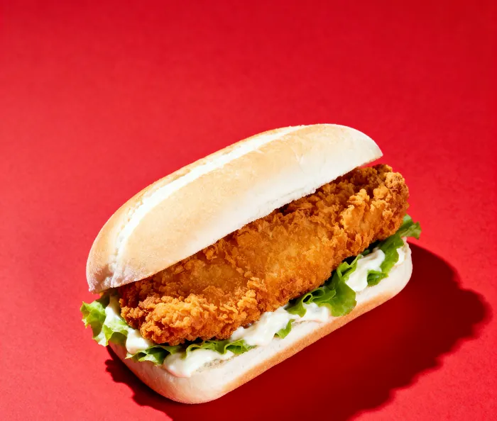 A crispy chicken sandwich on a soft white roll with lettuce and mayonnaise, on a red ground.