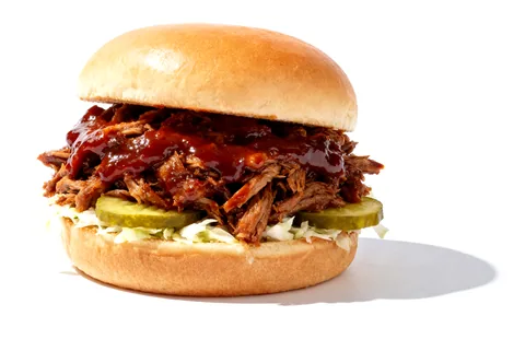 A pulled pork sandwich: a soft bun, pork in barbecue sauce, pickles and slaw.
